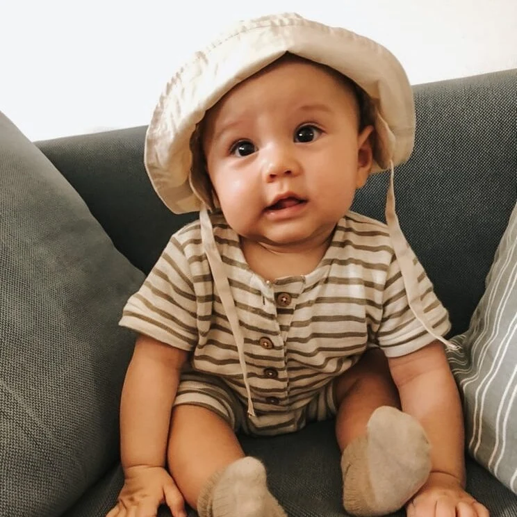 Blødt spedbarn i stribet tøj og sommerhat sidder på sofaen og kigger nysgerrigt.