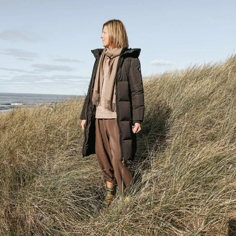 Udendørs kvinde i vinterfrakke på græsbevokset strandnært område, natur, kyst, hav, blåt himmel, vindfuldt vær.