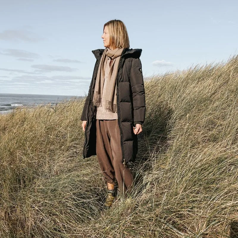 Udendørs kvinde i vinterfrakke på græsbevokset strandnært område, natur, kyst, hav, blåt himmel, vindfuldt vær.